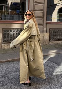 Vaaleahiuksinen nainen, jolla on päällään ylisuuri beige trench coat, mustat aurinkolasit ja mustat korkosaappaat, kävelee kaupungin kadulla ravintolan ikkunan lähellä.