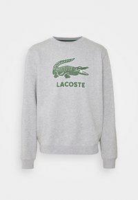Sudadera gris de manga larga, con un logo de cocodrilo verde bordado y el texto "LACOSTE" debajo. Puños y cuello acanalados.