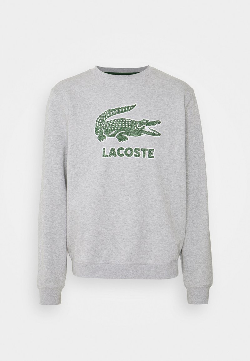 Sudadera gris de manga larga, con un logo de cocodrilo verde bordado y el texto "LACOSTE" debajo. Puños y cuello acanalados.