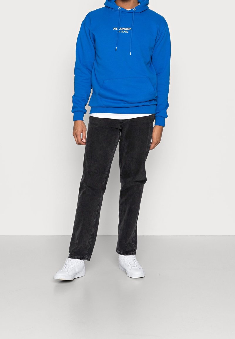 Felpa con cappuccio in cotone blu con testo stampato bianco e coulisse, abbinata a pantaloni in denim nero e sneakers bianche, posata su uno sfondo neutro.