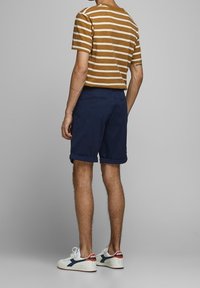 Raitapaita ruskeasta ja valkoisesta, lyhythihainen, tummansiniset käärityt shortsit, valkoiset tennarit sinisillä ja punaisilla yksityiskohdilla, nähtynä takaa.