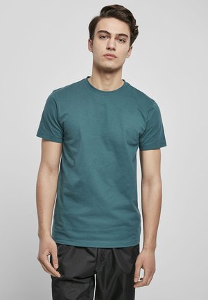 Teal cotton t-shirt with a crew neck and short sleeves, featuring a smooth texture and a relaxed fit. Worn with black trousers.