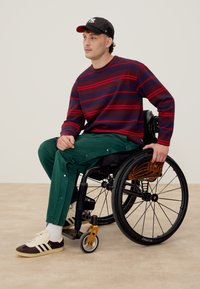 Férfi ül egy kerekesszékben, fekete-piros csíkos pulóvert, gombos zöld nadrágot, fehér zoknit, barna tornacipőt és fekete sapkát visel.