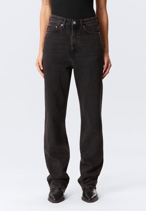 Person wearing high-waisted, loose-fit black jeans and black pointed-toe boots, standing against a plain light background.