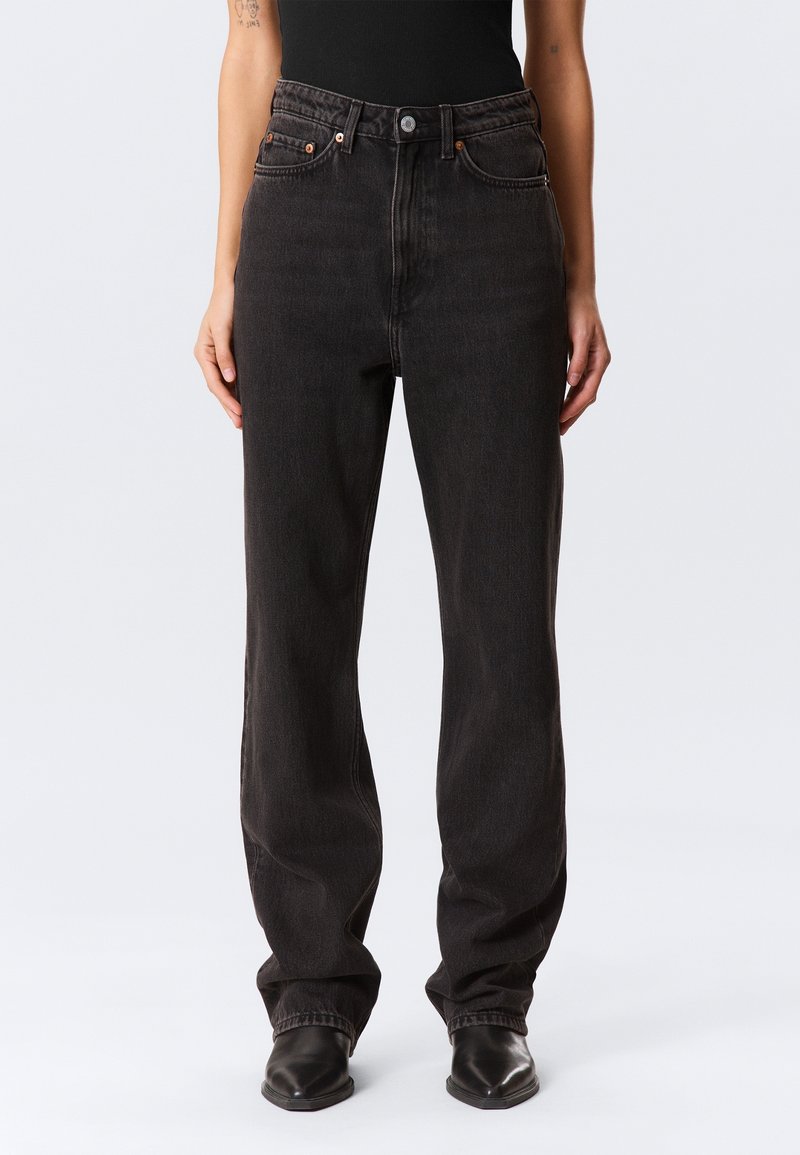 Personne portant un jean noir taille haute et ample, avec des bottes noires à bout pointu, se tenant devant un fond clair uni.