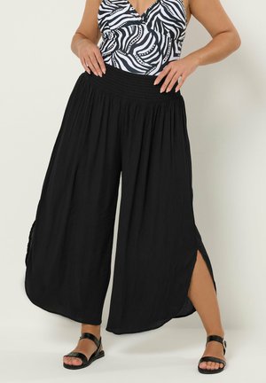 Kobieta ubrana w czarne spodnie z szerokimi nogawkami i rozcięciami po bokach, bezrękawnik w zebra wzór oraz czarne sandały na cienkich paskach na jednolitym tle.