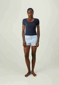 Ungdommelig svart kvinne som står barbeint iført en marineblå kortermet topp og lyseblå stripete shorts mot en ensfarget bakgrunn.
