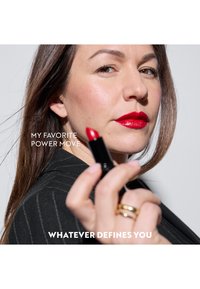 Rossetto rosso in un tubo nero tenuto da una donna con un blazer a righe sottili, capelli castani lisci e gioielli minimalisti. Sfondo bianco.