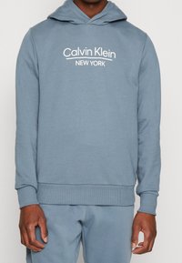Felpa leggera azzurro chiaro realizzata in morbido misto cotone, con tasca a marsupio, polsini e orlo in costina, e logo bianco "Calvin Klein New York".