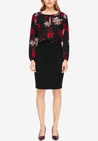 Femme portant une jupe crayon noire et un chemisier noir à manches longues avec des motifs floraux rouges et blancs, assortis à des escarpins bordeaux.