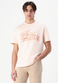 Camiseta de algodón rosa claro con mangas cortas que presenta un estampado gráfico de un cocodrilo y el texto "LACOSTE" en naranja. Ajuste casual.