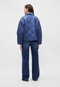 Γυναίκα που στέκεται με την πλάτη στραμμένη, φορώντας ένα μπλε επενδυμένο μπουφάν και φαρδιά σκούρα μπλε τζιν σε απλό φόντο.