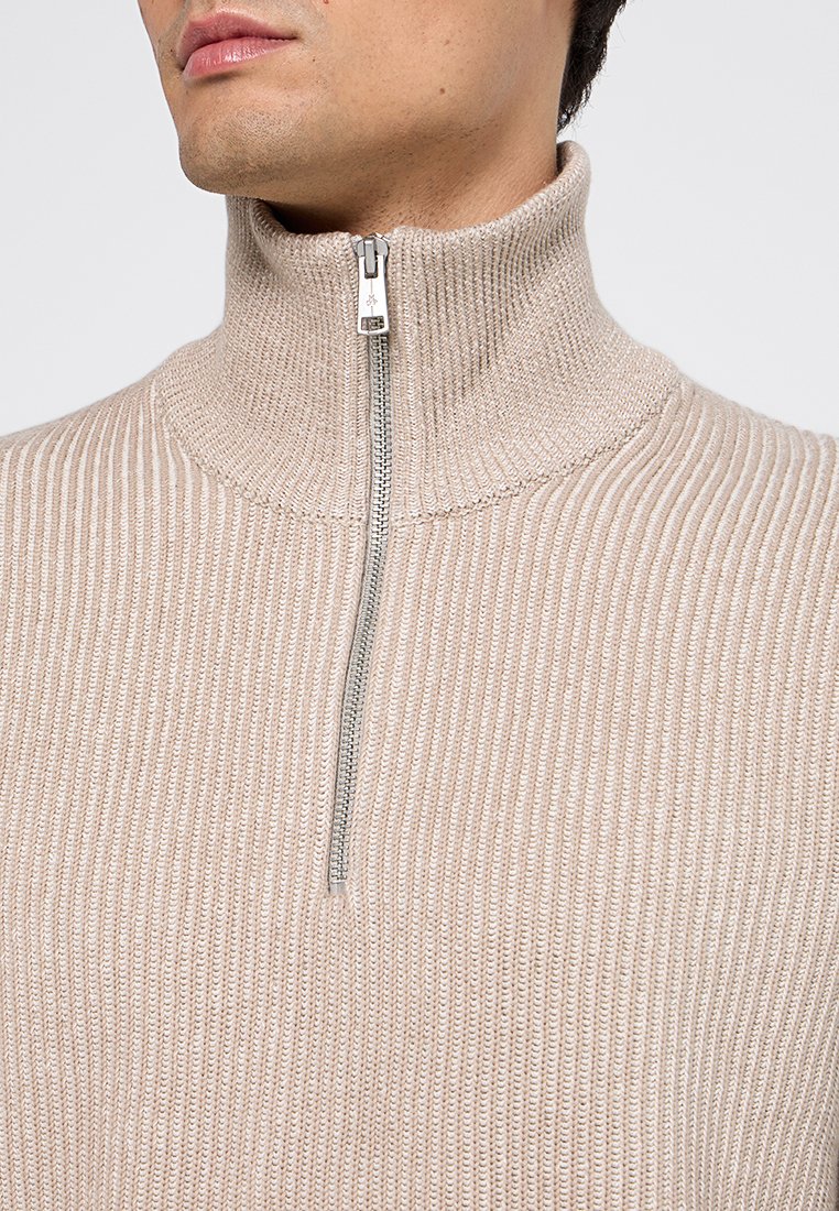 Mann trägt einen beigen, gerippten Strickpullover mit hohem Kragen und einem silbernen 1/2-Reißverschluss, Nahaufnahme auf Brust- und Halsbereich.