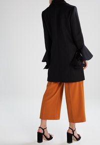 Sort uldfrakke med vidde ærmer, båret over orange vidde bukser. Sorte blokhælsandaler fuldender outfittet.