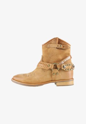 Bottine en daim camel avec un motif de couture au niveau de l'orteil, des sangles décoratives avec des clous métalliques, et un petit talon en bois sur fond blanc.