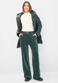 Grüne Pufferjacke über einem cremefarbenen Rollkragenpullover, kombiniert mit weit geschnittenen, samtartigen, dunkelgrünen Hosen und schwarzen klobigen Schuhen. Ein schwarzer Gürtel betont die Taille.