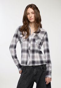 Camicia a quadri grigia e bianca con maniche lunghe, dotata di due tasche sul petto e un orlo leggermente accorciato, abbinata a jeans neri.