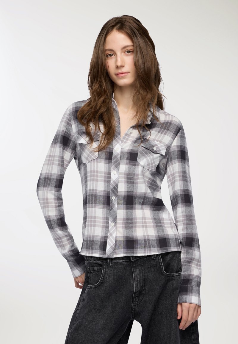 Camicia a quadri grigia e bianca con maniche lunghe, dotata di due tasche sul petto e un orlo leggermente accorciato, abbinata a jeans neri.