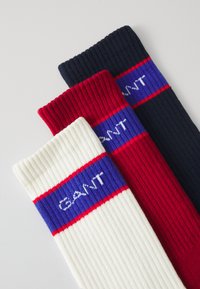 Drie geribde sokken in het wit, rood en marineblauw, elk met een blauwe band voorzien van het logo "GANT" in witte letters.