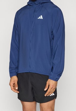 Homme portant une veste à capuche bleue marine zippée et un short noir, tous deux arborant un logo triangulaire blanc sur le côté gauche.