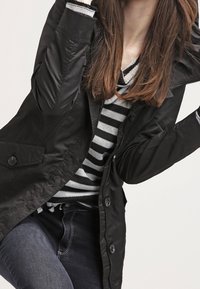 Svart, långärmad jacka med glansig yta, två stora framslutande knappar och figurnära design som bärs över en randig tröja och mörka jeans.