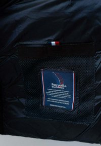 Gros plan sur une poche de veste noire avec une étiquette bleue aux couleurs du drapeau français et du texte sur la Patrouille de France par la collection Redskins.