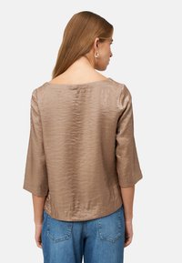 Braune metallische Bluse mit lockerer Passform, weitem Ausschnitt und dreiviertelärmeln, kombiniert mit blauen High-Waist-Jeans. Glatte Textur.