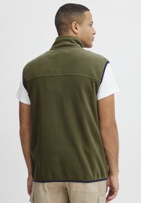 Fleece vest i olivengrønn med høy krage, med marineblå kant langs armhulene og en glatt tekstur. Vist fra baksiden.