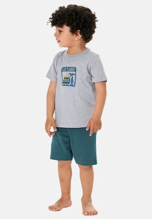 Young boy with curly hair wearing gray "Go Surf" t-shirt and teal shorts, standing barefoot and looking to the side against a white background.