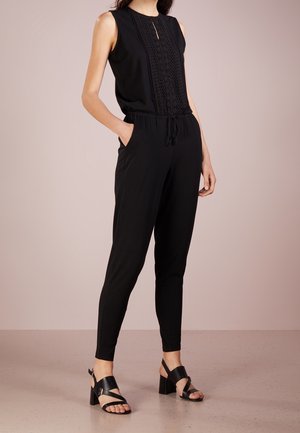 Vrouw draagt een mouwloze zwarte jumpsuit met kantdetails en een koord in de taille, gecombineerd met zwarte sandalen met hakken, staat met handen in de zakken.