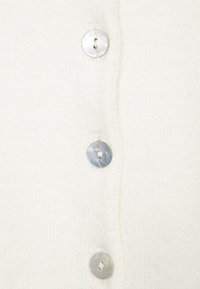 Vit stickad kofta med sex runda, glänsande silverknappar och synlig texturerad sömnad längs placket.