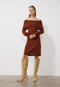 Off-the-shoulder, rostfärgad stickad klänning med långa ärmar, en figurnära silhuett och framhävd ribbad halsringning, kombinerad med beige knähöga stövlar.