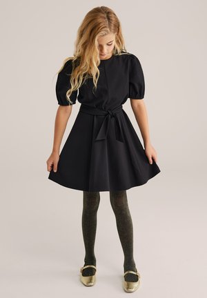 Fekete, rövid ujjú ruha övvel a deréknál, bővülő szoknyával, sima textúrával, csillogó arany balettcipőkkel és csillogó fekete harisnyával párosítva.