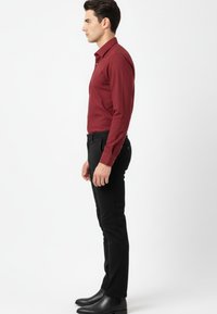 Hombre de pie de perfil vistiendo una camisa ajustada de manga larga burdeos, pantalones slim negros y zapatos de vestir negros, contra un fondo blanco.