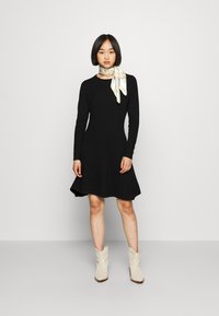Vestido preto de mangas compridas com uma bainha rodada. Combinado com um lenço estampado em branco e dourado. Usado com botins bege.