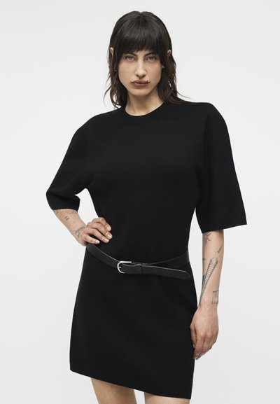 Γυναίκα με σκούρα μαλλιά που φοράει μαύρο φόρεμα κοντού μανικιού με ζώνη, ποζάρει με το ένα χέρι στη μέση, δείχνοντας τα τατουάζ στο χέρι.