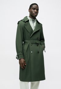 Trench coat verde doppiopetto, con mostrine sulle spalle, cintura in vita e grandi bottoni, indossato sopra una camicia a righe.