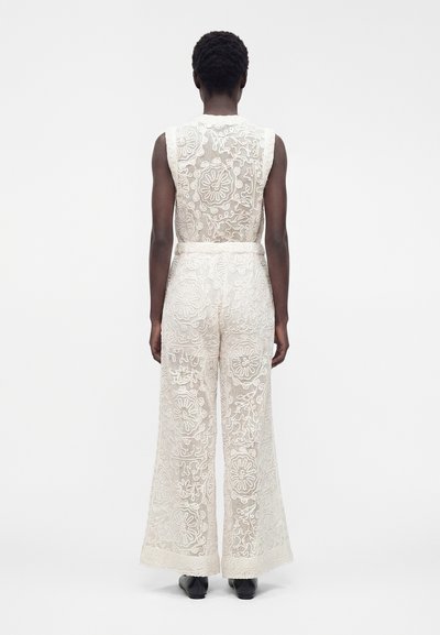 Personne noire debout, portant un haut en dentelle blanche sans manches et un pantalon large en dentelle à motifs floraux, face à un fond blanc uni.