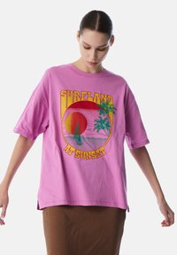 T-shirt oversize rosa chiaro con una grafica che rappresenta un tramonto, palme e una barca. Maniche corte, in cotone, vestibilità casual.