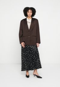 Brūns garš žakete ar vienu pogu, kombinēts ar plūstošu melnu svārku ar baltām pumpiņām un melniem, punktainiem papēžiem.