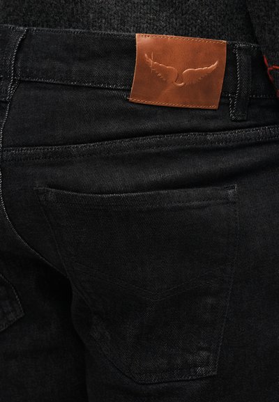 Černé džíny z denimu mají hladkou texturu, jemný lesk a na pásku je kožená záplata s reliéfním motivem křídel. Je viditelná zadní kapsa.