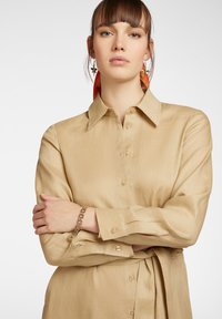 Camicia beige con bottoni, realizzata in un tessuto morbido, con maniche lunghe, colletto, bottoni frontali e dettaglio in vita legato. Presenta un bracciale dorato.