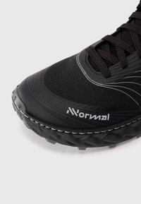 Chaussure de sport noire avec une tige texturée, un embout renforcé et une semelle en caoutchouc avec des rainures profondes pour une meilleure traction. Accents réfléchissants présents.