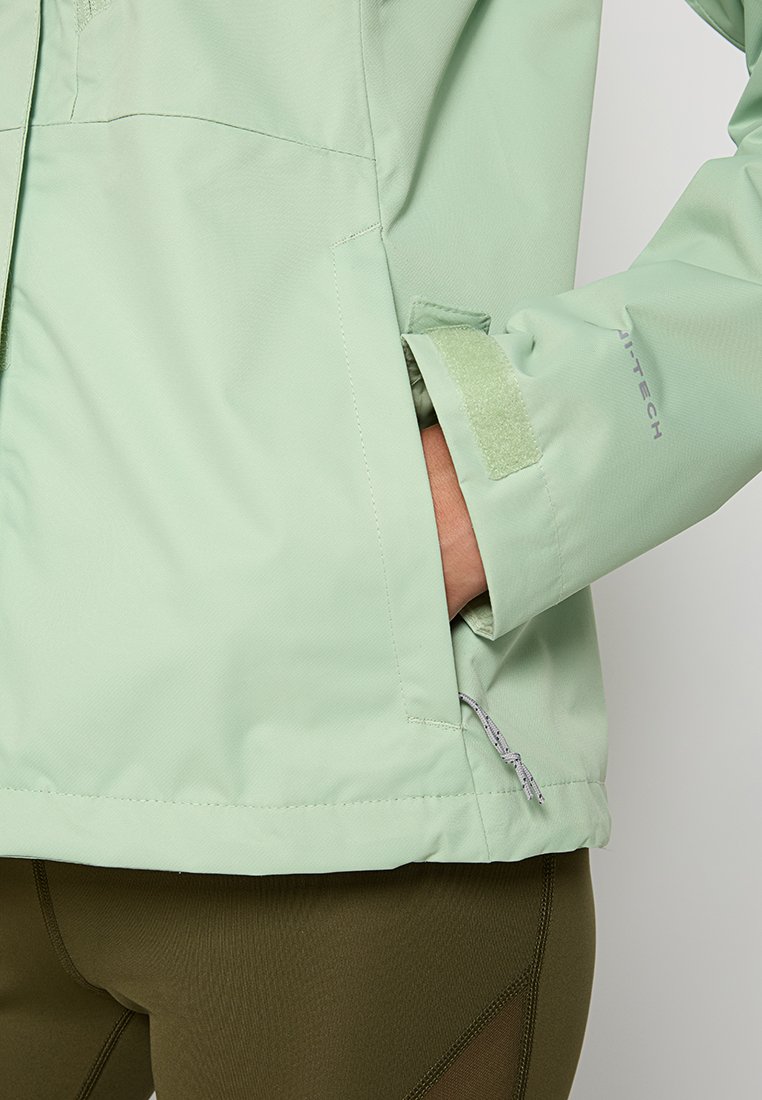 Veste imperméable vert clair avec une poche zippée, une fermeture à patte auto-agrippante aux poignets et un tissu texturé, associée à un pantalon vert olive.