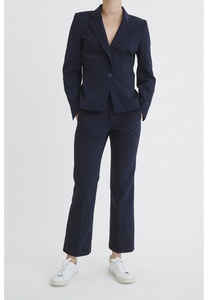 Person trägt einen maßgeschneiderten marineblauen Anzug mit einem einreihigen Sakko und passenden Hosen, kombiniert mit weißen Sneakers, steht mit den Händen in den Taschen.