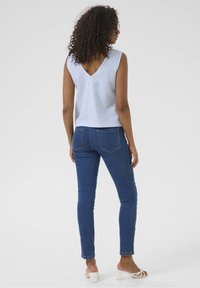 Donna con capelli ricci, che indossa un top a righe senza maniche, jeans blu aderenti e sandali con tacco bianchi, vista di spalle su uno sfondo neutro.