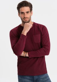 Burgundy pitkähihainen paita, jossa on teksturoitu pinta, pyöreä kaula-aukko ja rintatasku. Pehmeä kangas ja istuva malli.