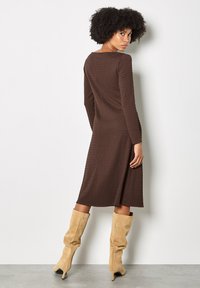 Brown, ribbed long-sleeve dress with a flared silhouette, mid-calf length, and a square neckline, paired with knee-high beige boots.