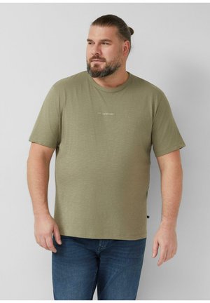 Bärtiger Mann mit zurückgebundenem Haar, der ein olivgrünes T-Shirt mit dem Text "LET'S SYNERGIZE!" und blaue Jeans trägt und vor einem schlichten hellen Hintergrund steht.