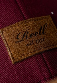 Etiqueta de cuero marrón con texto en relieve "Riell est. 1997" sobre un fondo de tejido texturizado color burdeos, con costuras cuidadosas a lo largo de los bordes.